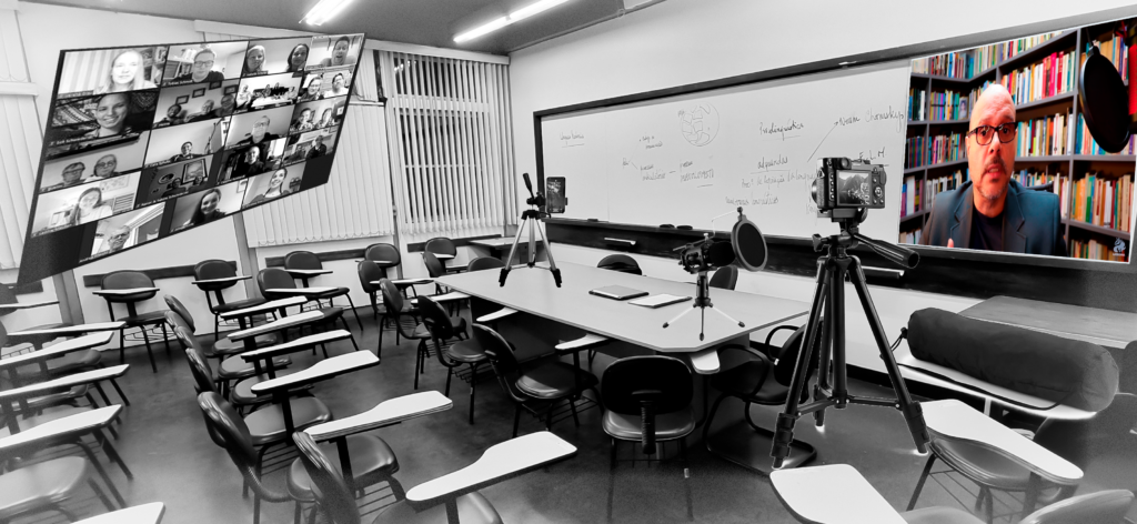 Figure - Classroom figuring teacher's table, universitary armchairs and whiteboard includes elements that indicate the hyperspecialty of the class: floating screen with images of online students cameras; tele-broadcast equipment (low tripod cell phone camera on the table; high tripod camera on the floor; microphone and TV screen on the whiteboard with real-time transmitted image)